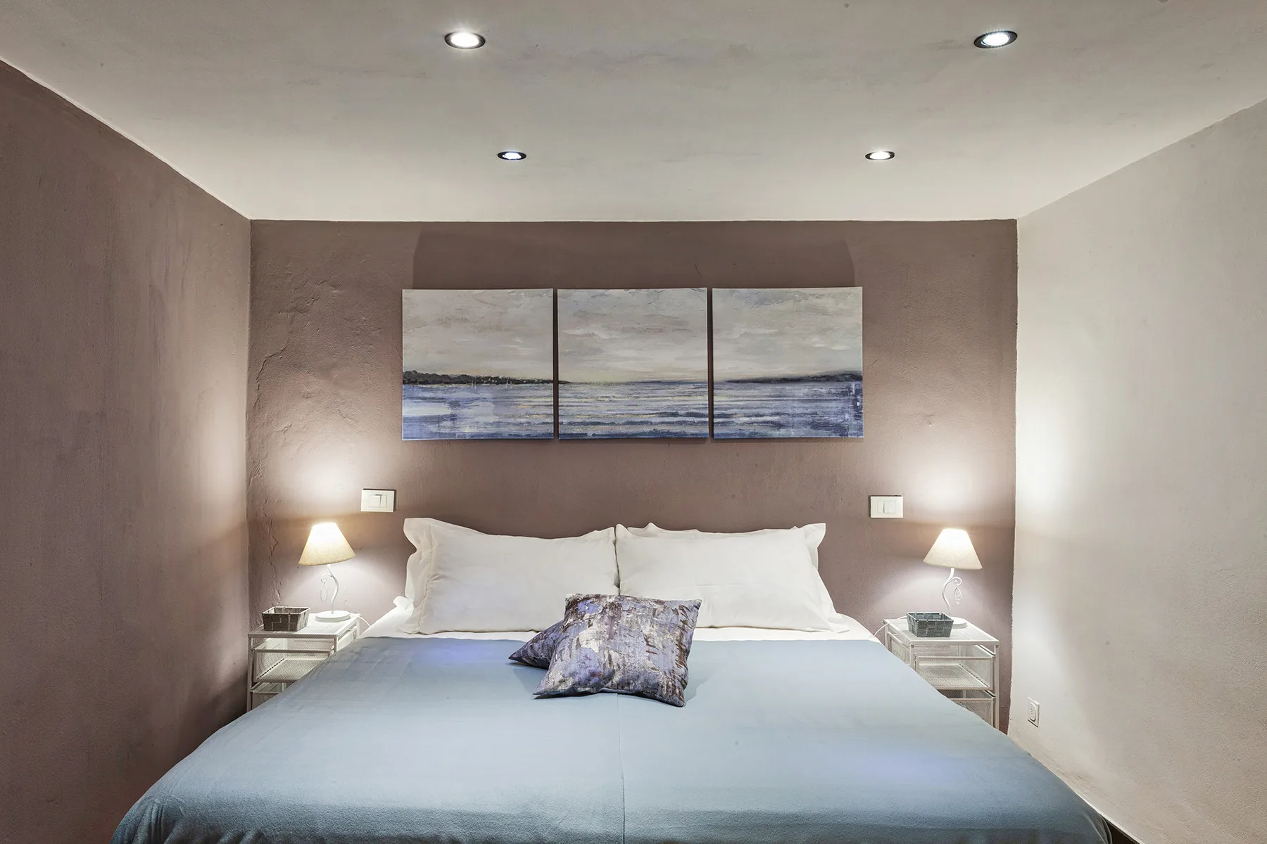 Un dormitorio con una cama bien tendida y un cuadro de paisaje marino en tres paneles.