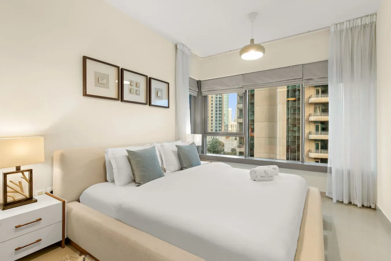 A bedroom with a neatly made bed beside large windows overlooking nearby buildings.