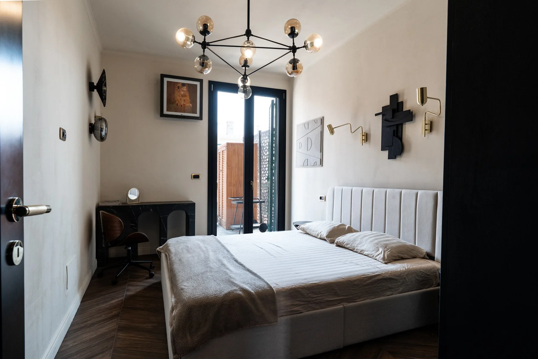 A bedroom with a large bed, a desk, and glass doors opening to a balcony.