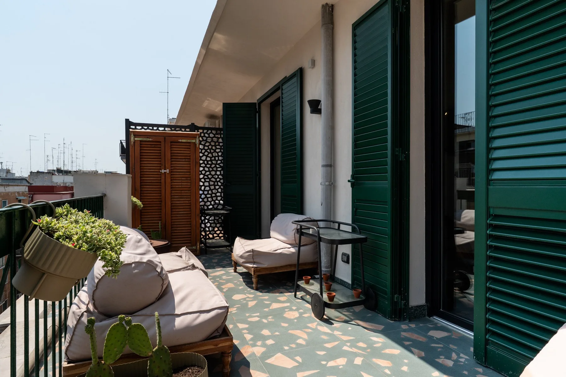 A sunny outdoor terrace has cushioned seating, potted plants, and dark green shutters.