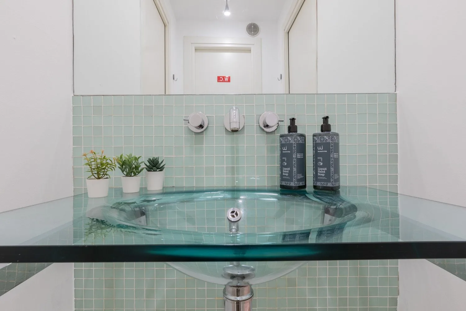 A tiled bathroom vanity with a glass countertop, basin, and small potted plants.