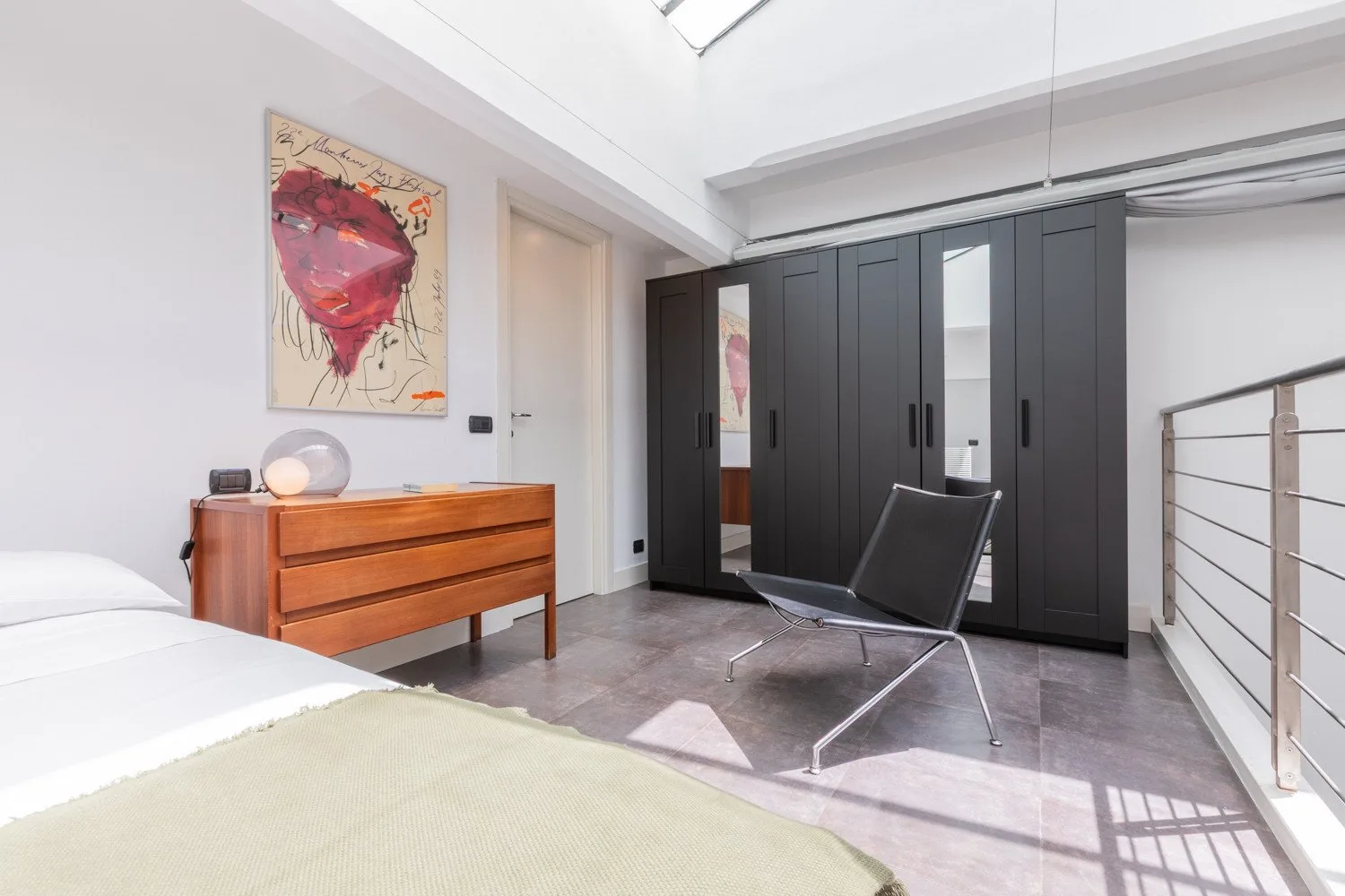 A bright bedroom with a bed, wooden dresser, and dark built-in wardrobes.