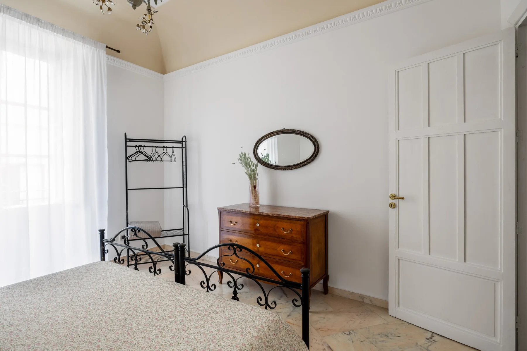 A bedroom with an iron bed frame, wooden dresser, and oval wall mirror.
