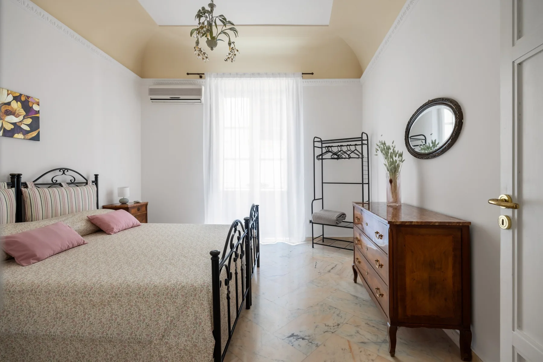 A bedroom with a bed, a dresser, and a tall window with sheer curtains.