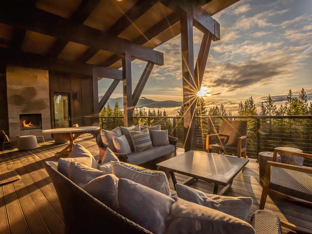 Una sala de estar al aire libre con asientos y una mesa mira hacia un paisaje boscoso al atardecer.