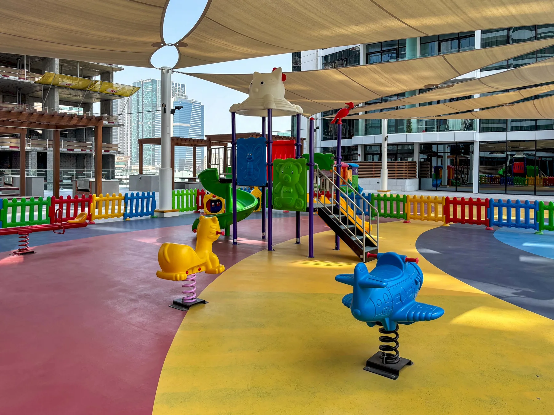 Un colorido parque infantil al aire libre con juegos para trepar y balancines bajo toldos.