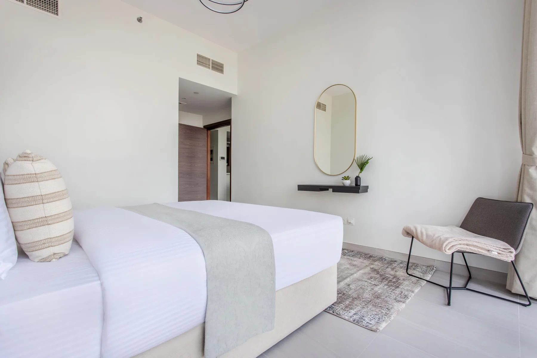 Un dormitorio luminoso con una cama tendida, un espejo ovalado y una silla pequeña.