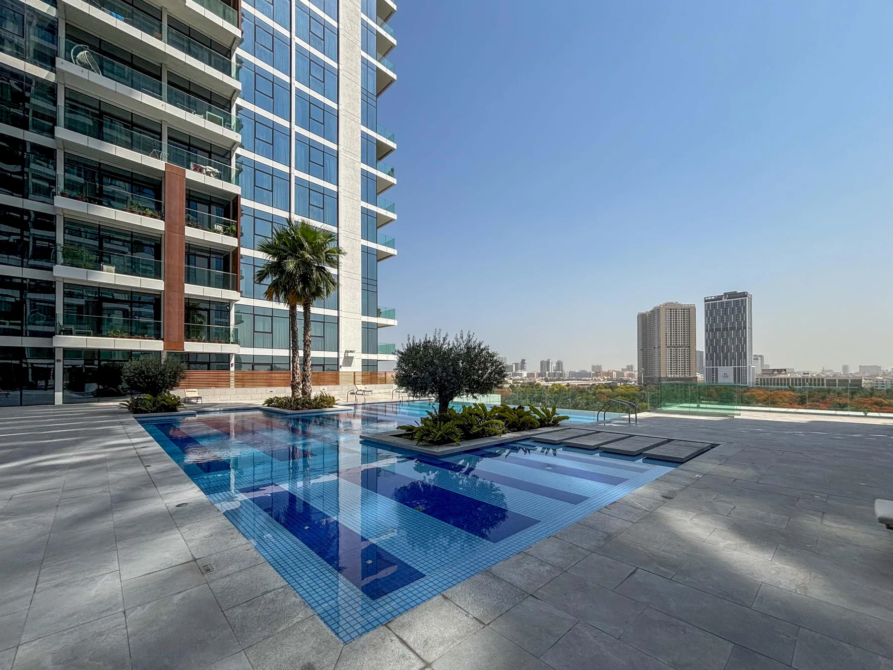 Una piscina al aire libre junto a un edificio alto moderno, con edificios urbanos a lo lejos.