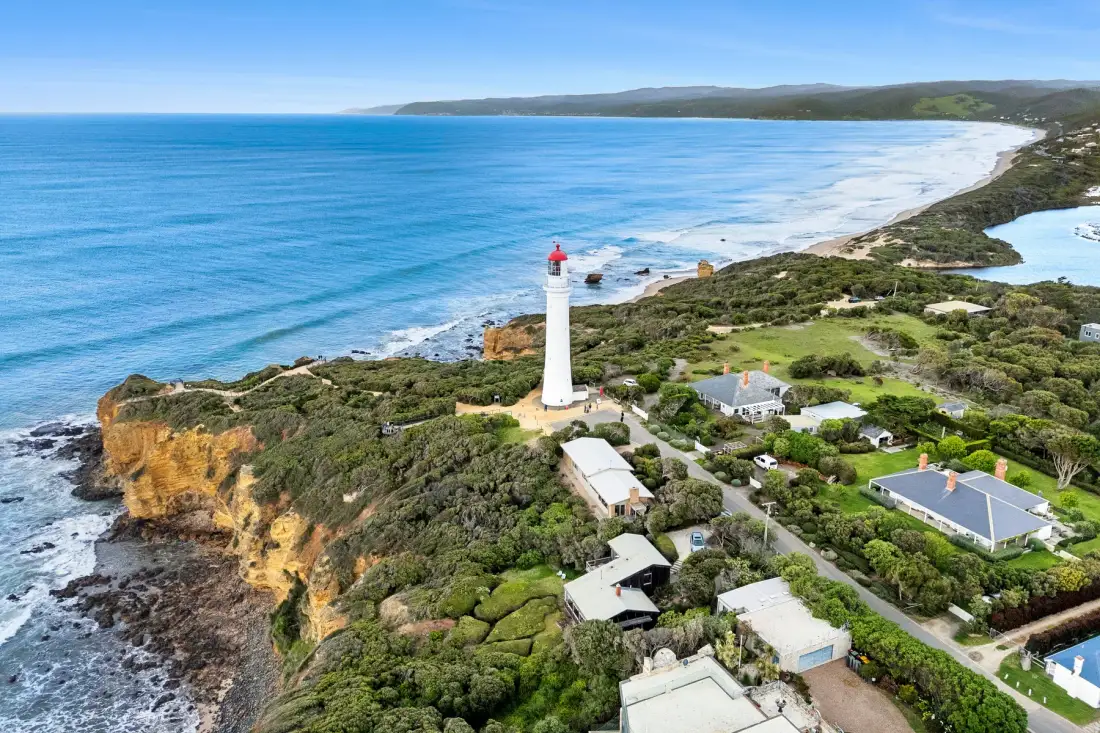 Aerial coastal scene with a lighthouse, ocean, and nearby homes.
