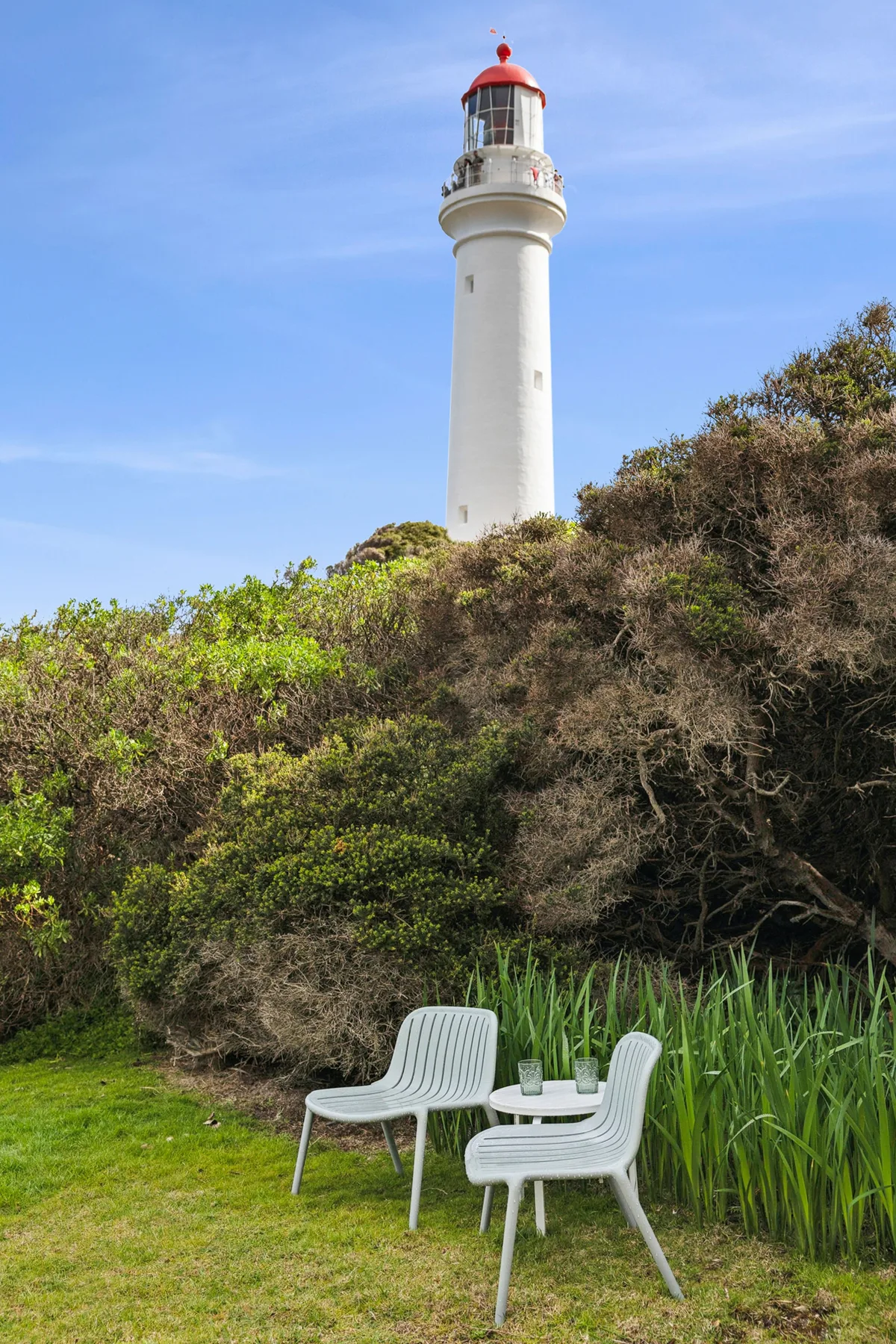A white lighthouse rises above a green hillside beside outdoor seating.