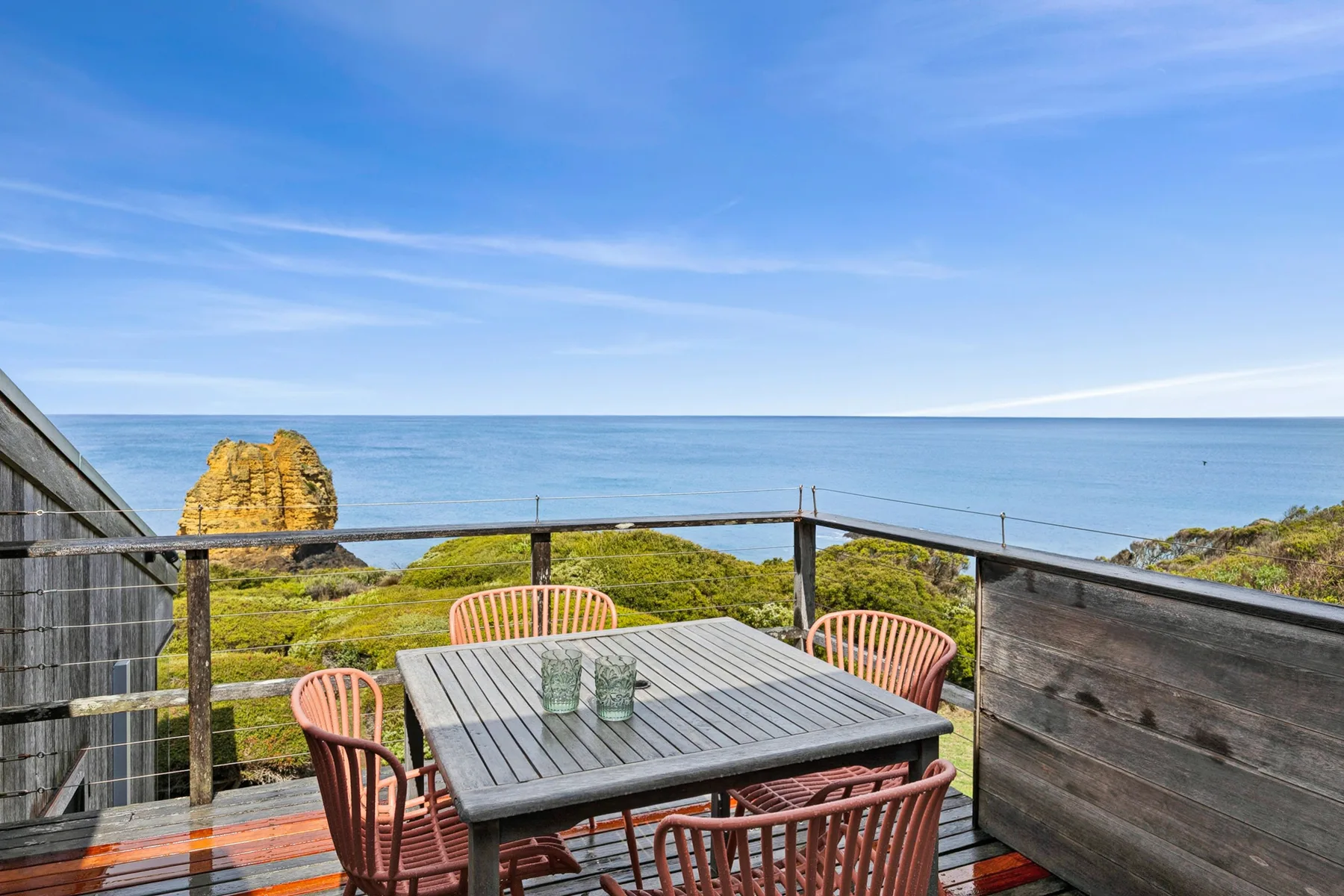 An outdoor dining area overlooks the ocean, with a table and seating on a wooden deck.