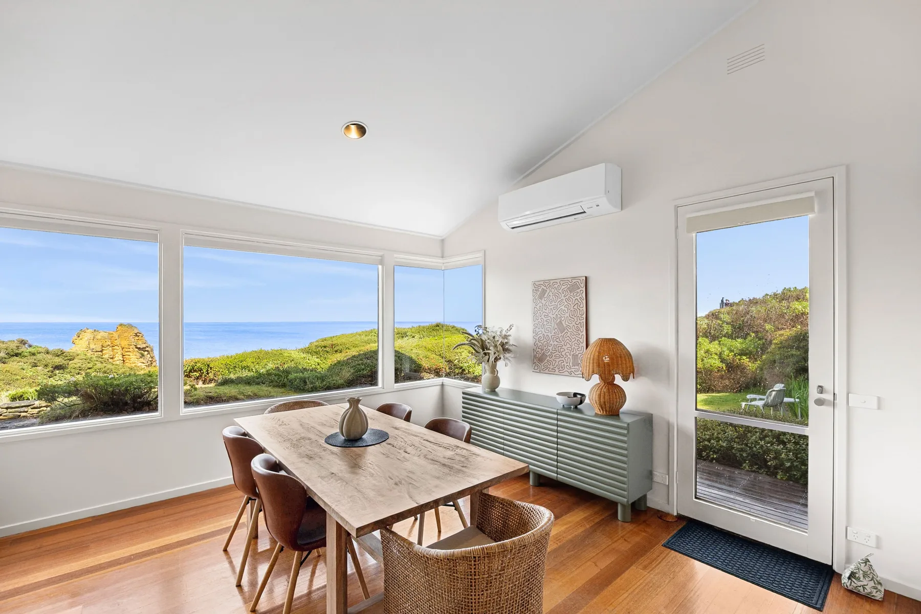A bright dining area with a long table beside wide windows overlooking the ocean.