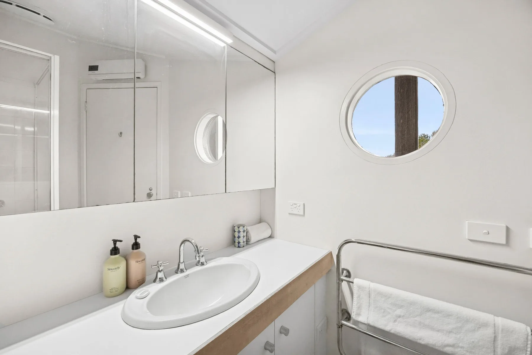 A bright bathroom with a sink, mirrored cabinets, and a round window.