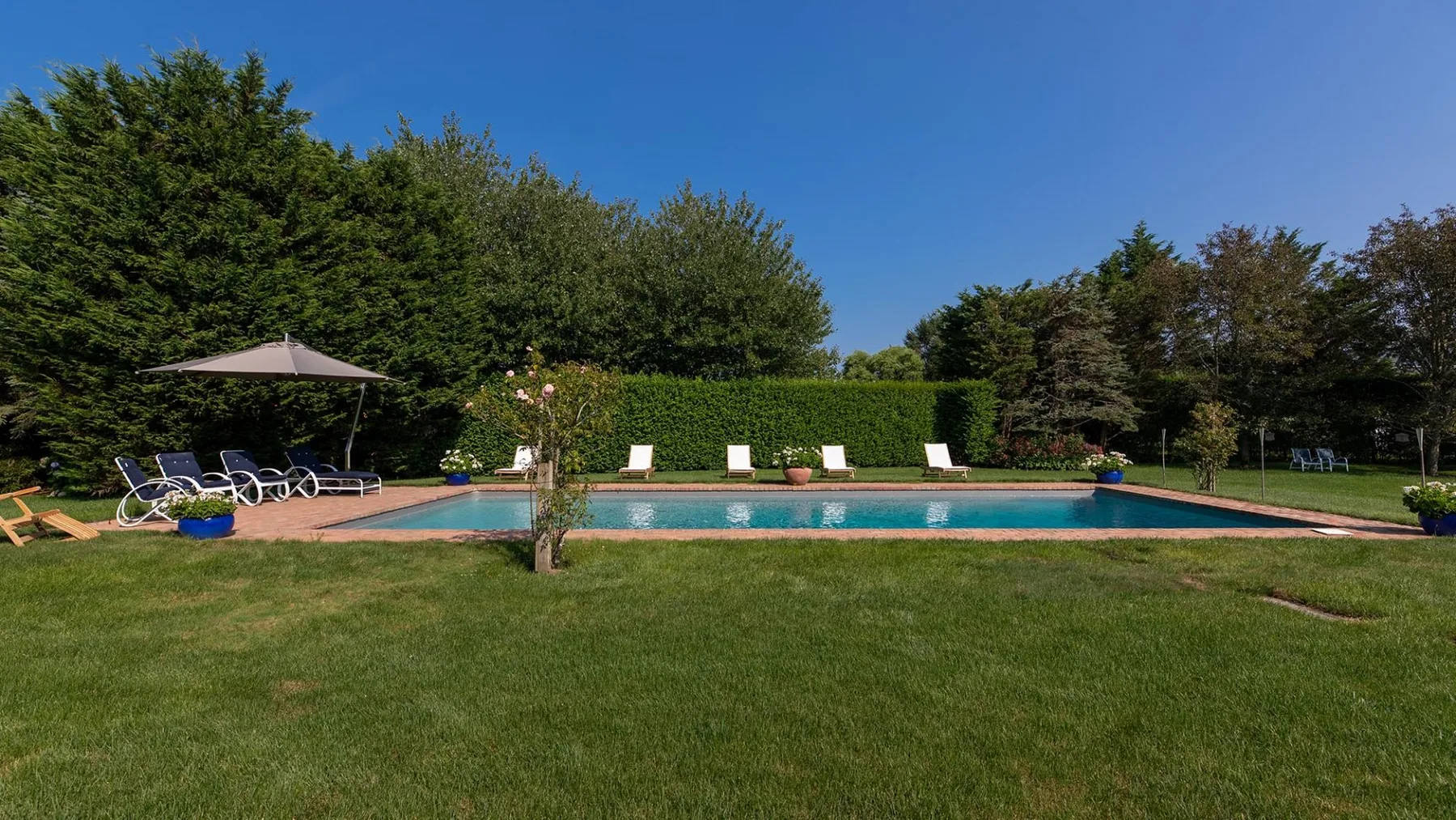 Una piscina al aire libre está junto a un amplio césped, con asientos y árboles alrededor.