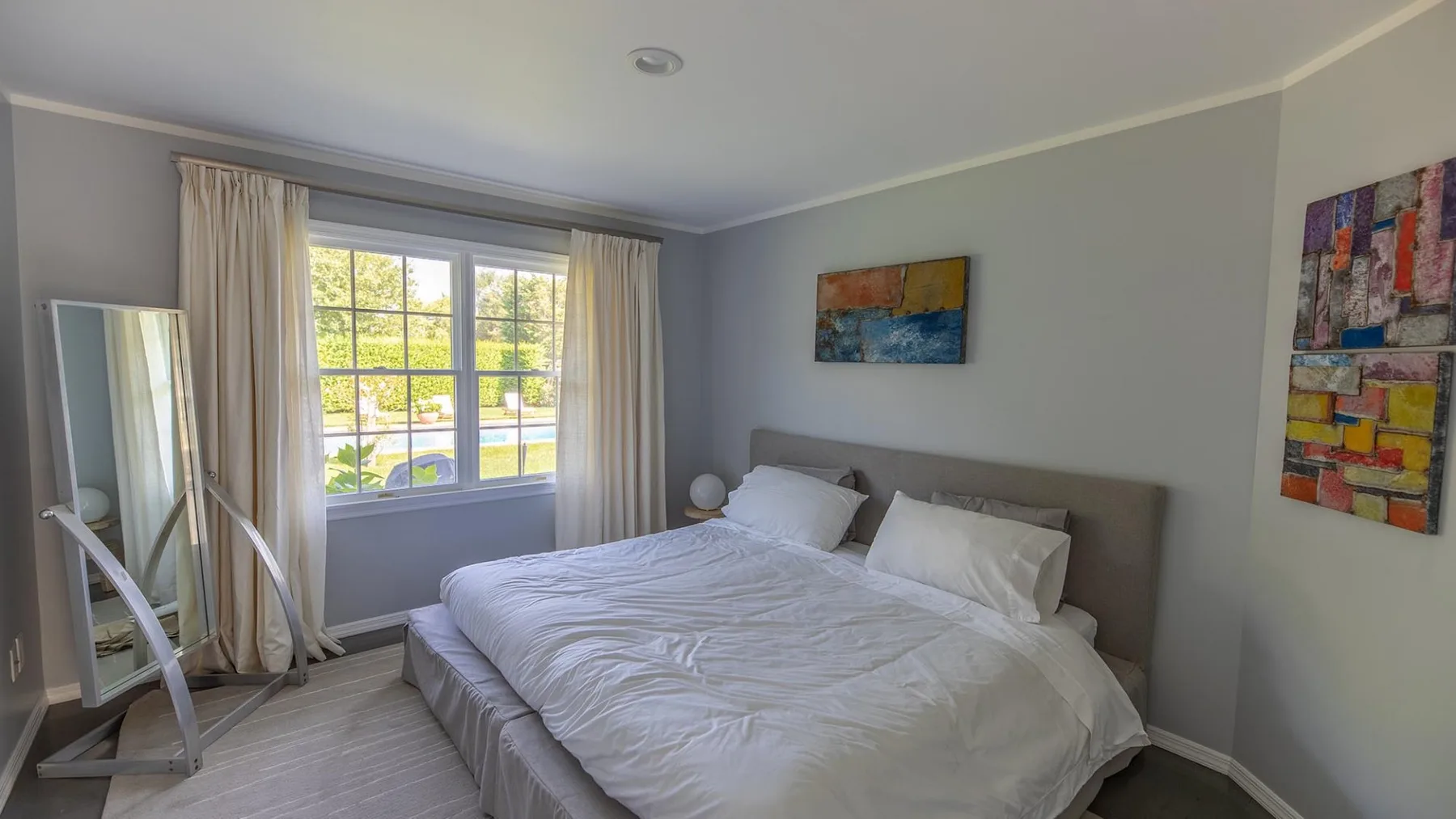 Un dormitorio con una cama junto a una ventana grande y cuadros coloridos.
