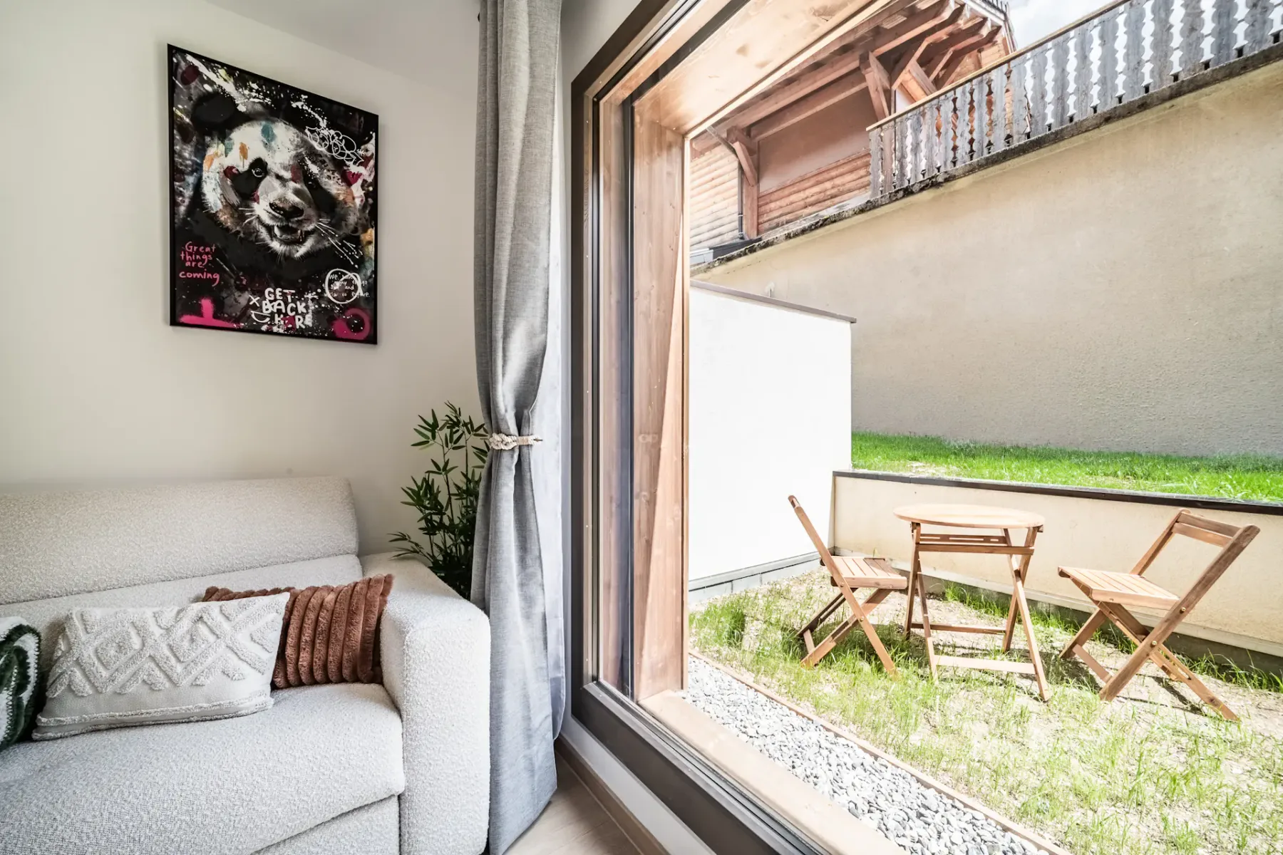 Cozy living room with a white sofa, decorative cushions, and art. Large window overlooks a small garden with a wooden table and chairs.