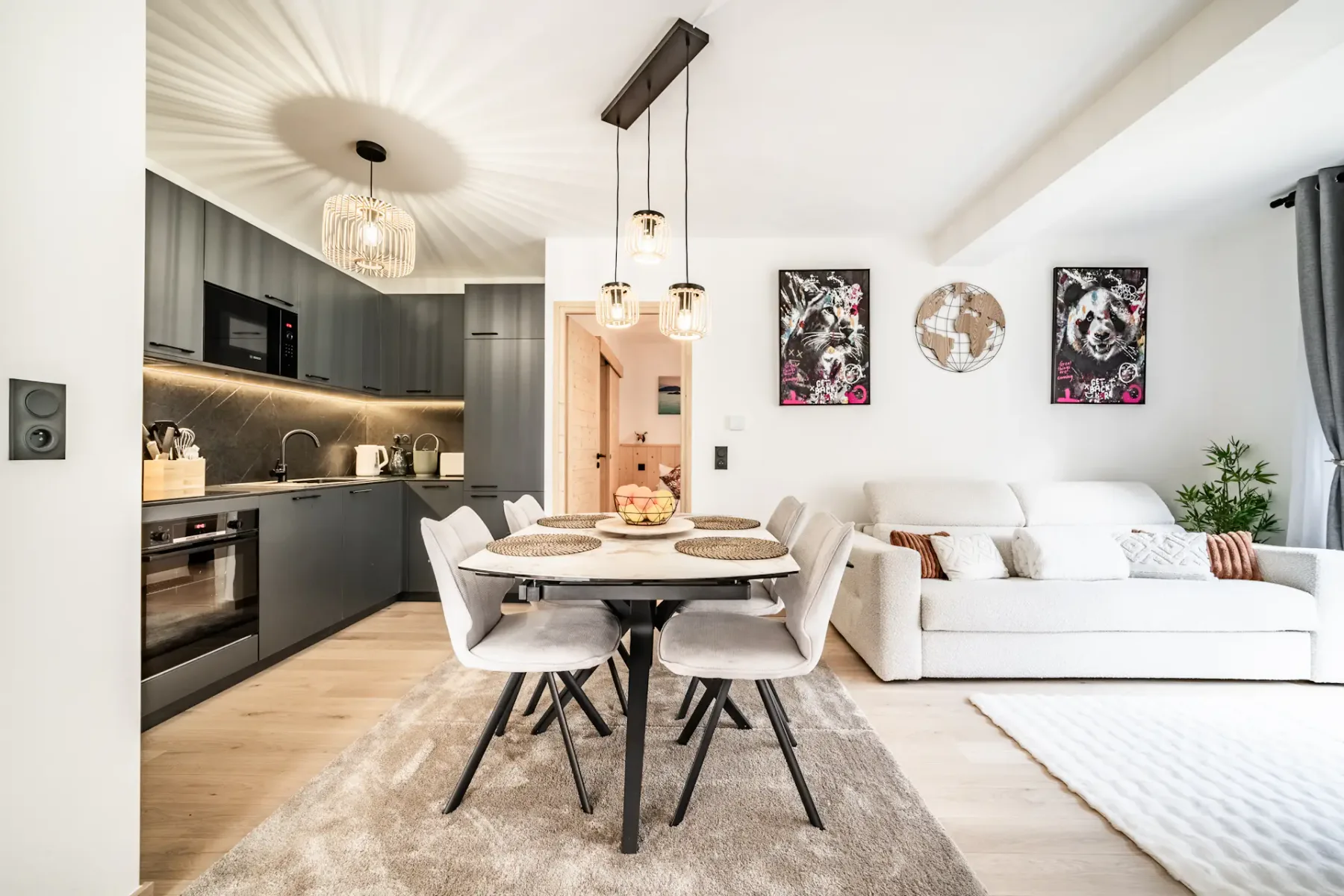 Modern living and dining area with light furnishings, a table set for four, pendant lights, artwork, and glass doors leading to a garden patio.