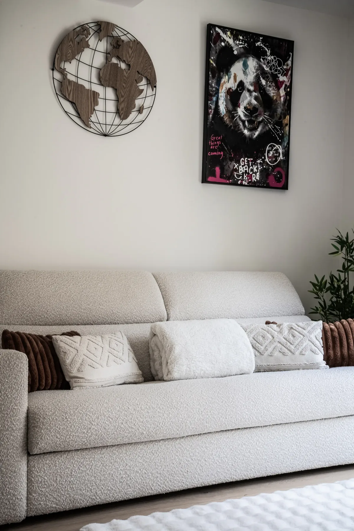 Cozy living room with a white textured sofa, patterned pillows, a metal world map, a colorful panda picture, and a small plant in the corner.