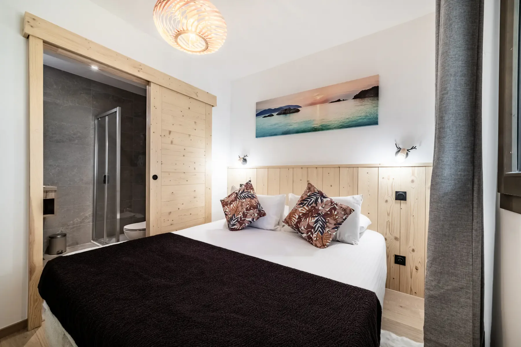 Cozy bedroom with a double bed, decorative pillows, wooden accents, a framed landscape photo, modern lighting, and a window with curtains.
