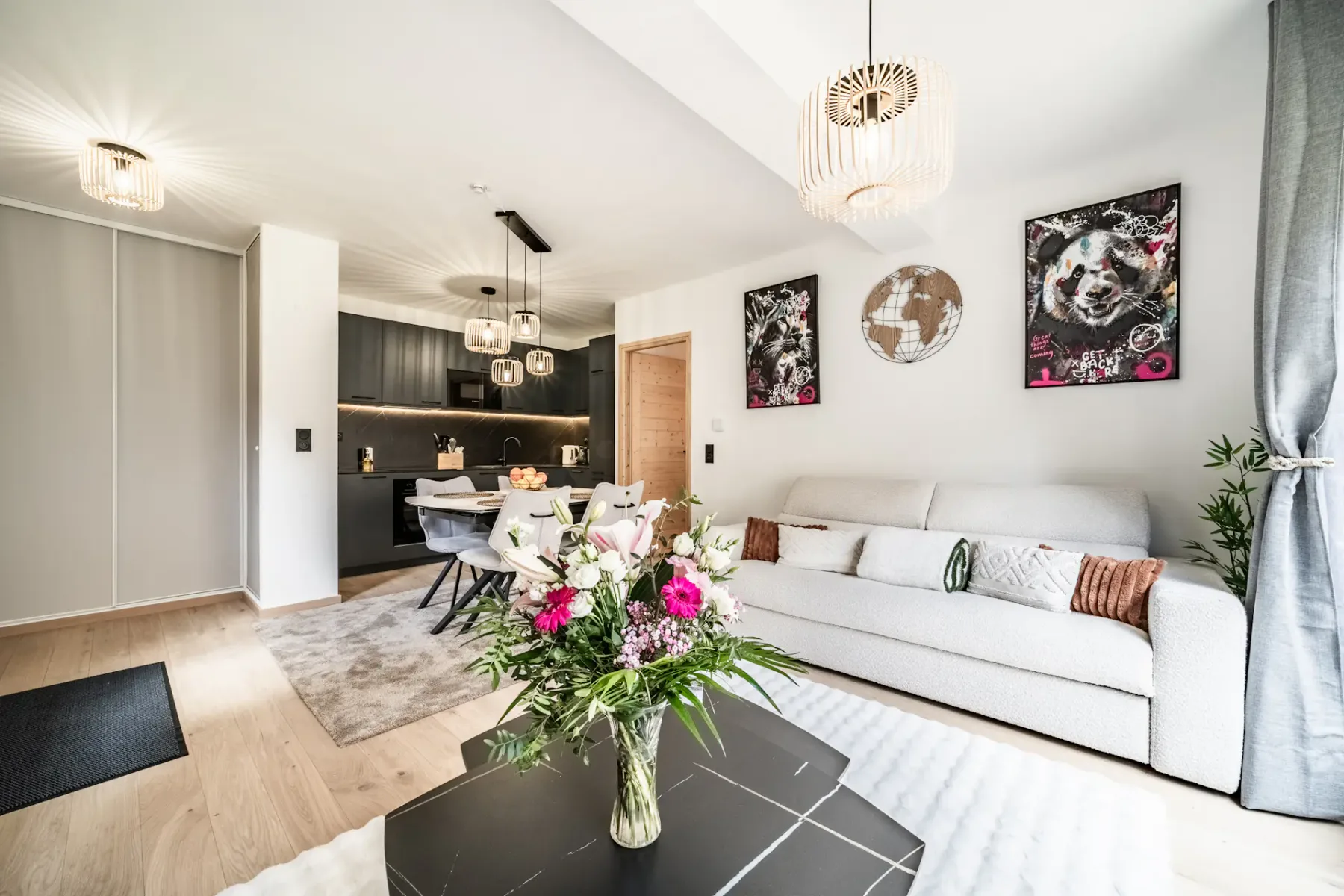 Bright living room with a white sofa, black coffee table, floral centerpiece, art on the walls, large windows, and gray curtains.