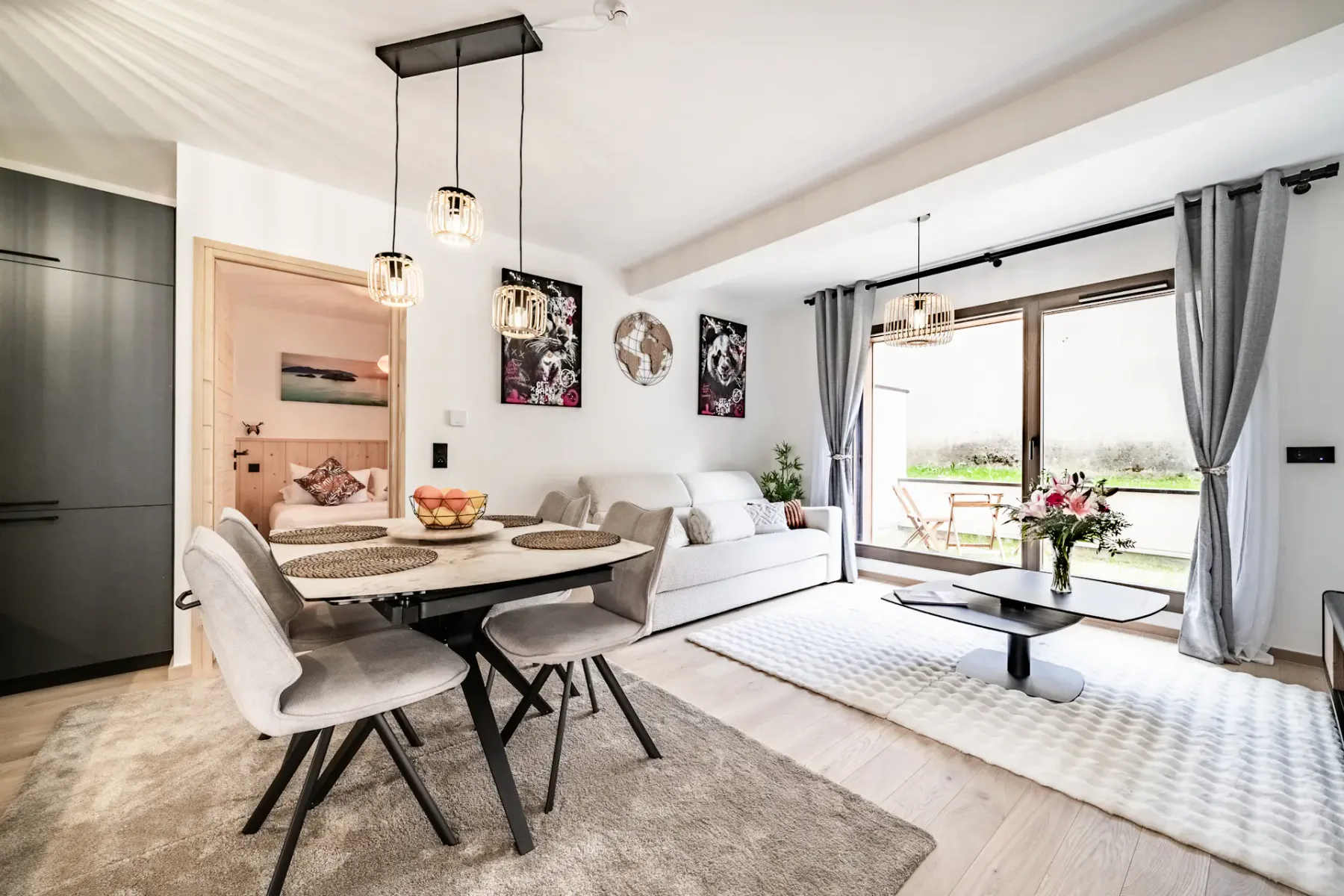 Modern living and dining area with light furnishings, a table set for four, pendant lights, artwork, and glass doors leading to a garden patio.