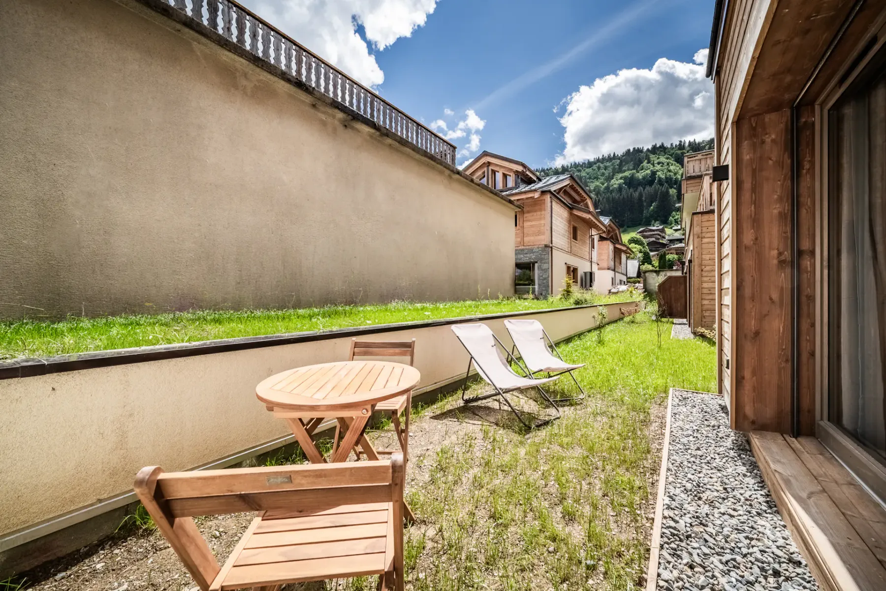 Modern building courtyard with greenery, wooden accents, open door, and mountain view under a partly cloudy blue sky.