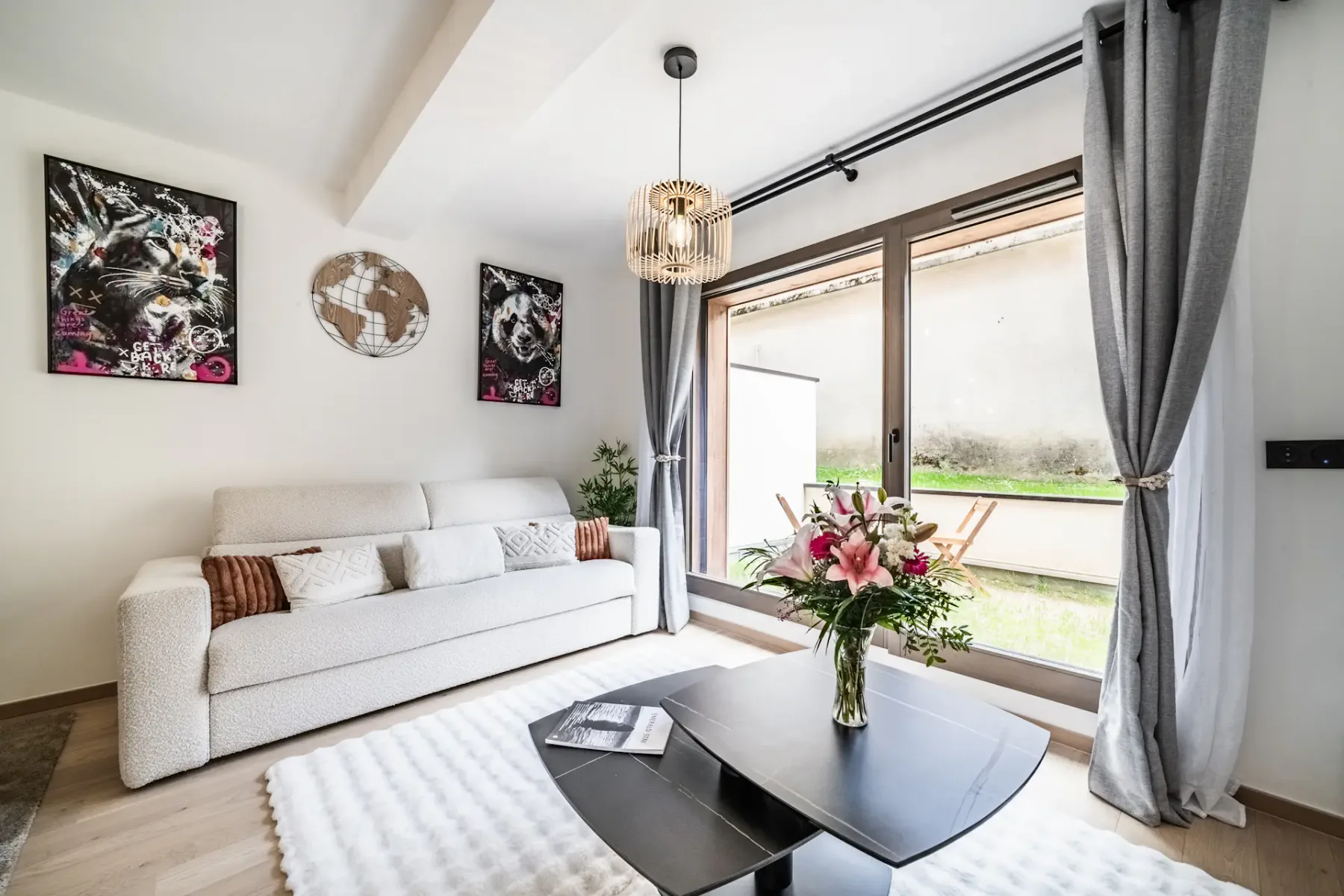 Bright living room with a white sofa, black coffee table, floral centerpiece, art on the walls, large windows, and gray curtains.