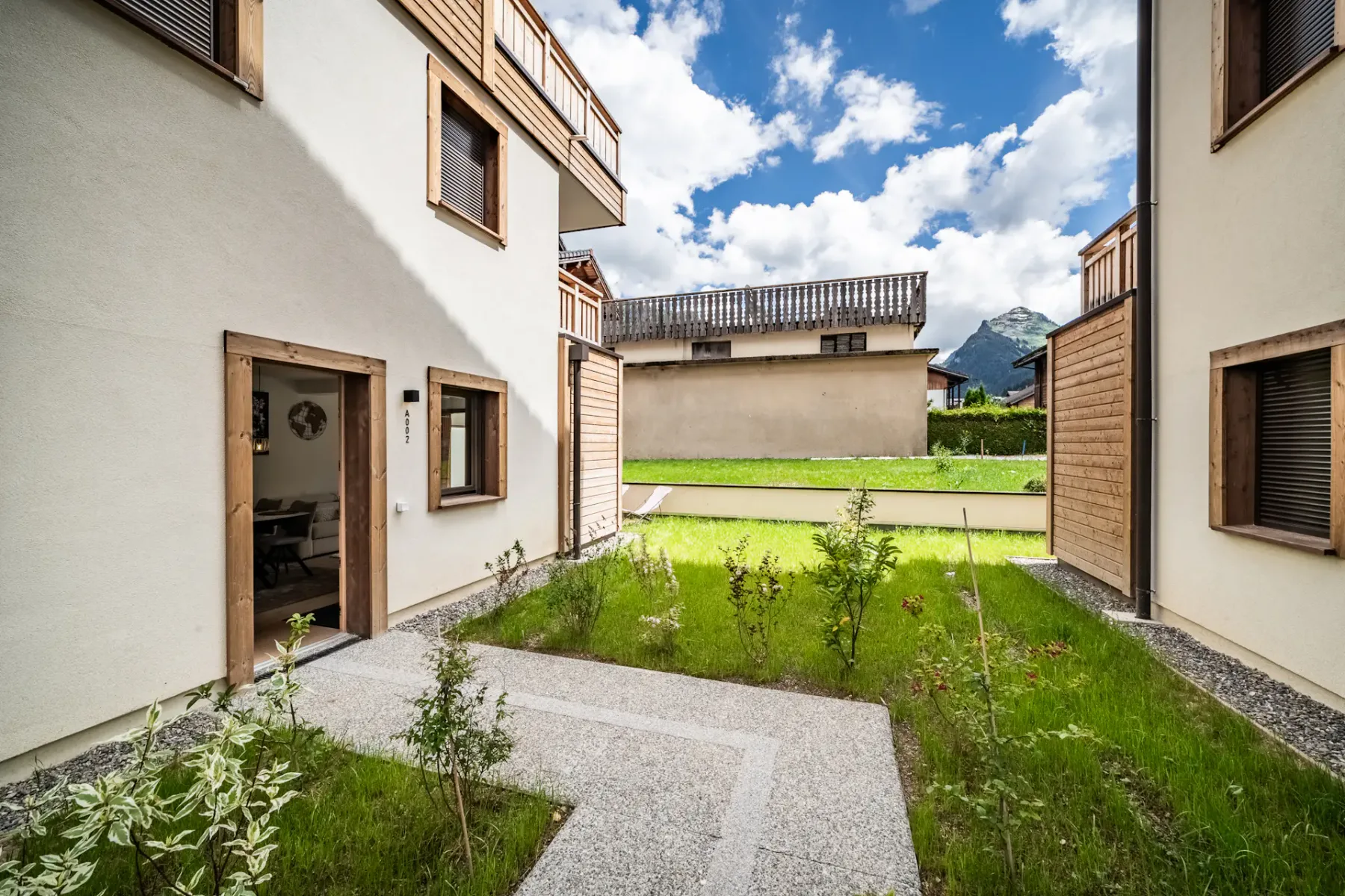Modern building courtyard with greenery, wooden accents, open door, and mountain view under a partly cloudy blue sky.