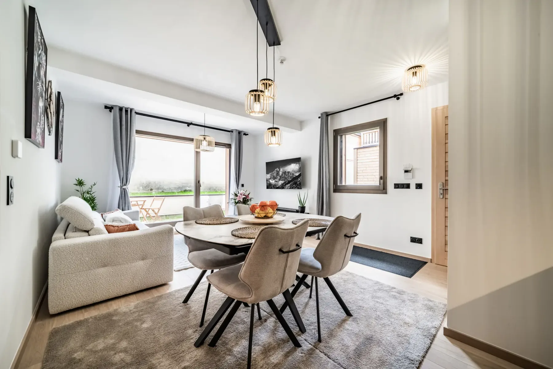 Modern living and dining area with light furnishings, a table set for four, pendant lights, artwork, and glass doors leading to a garden patio.