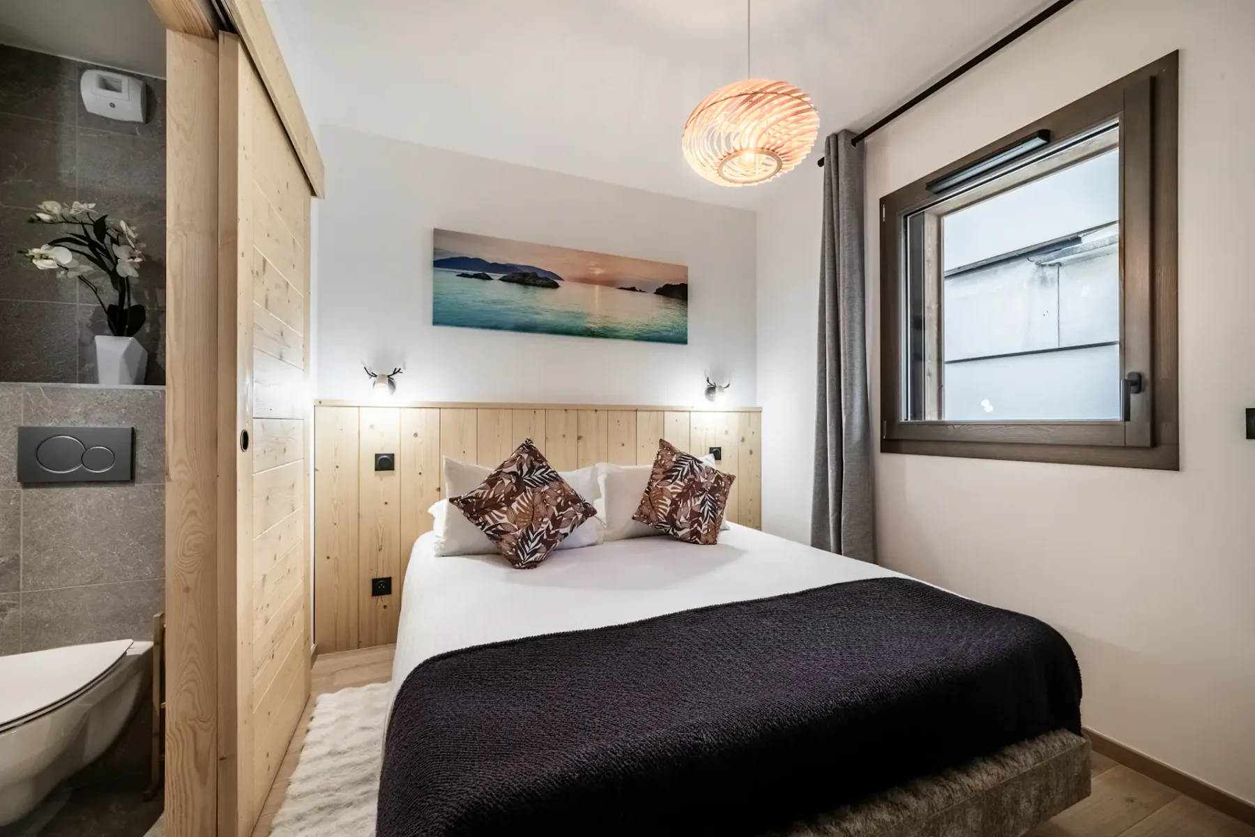 Cozy bedroom with a double bed, decorative pillows, wooden accents, a framed landscape photo, modern lighting, and a window with curtains.