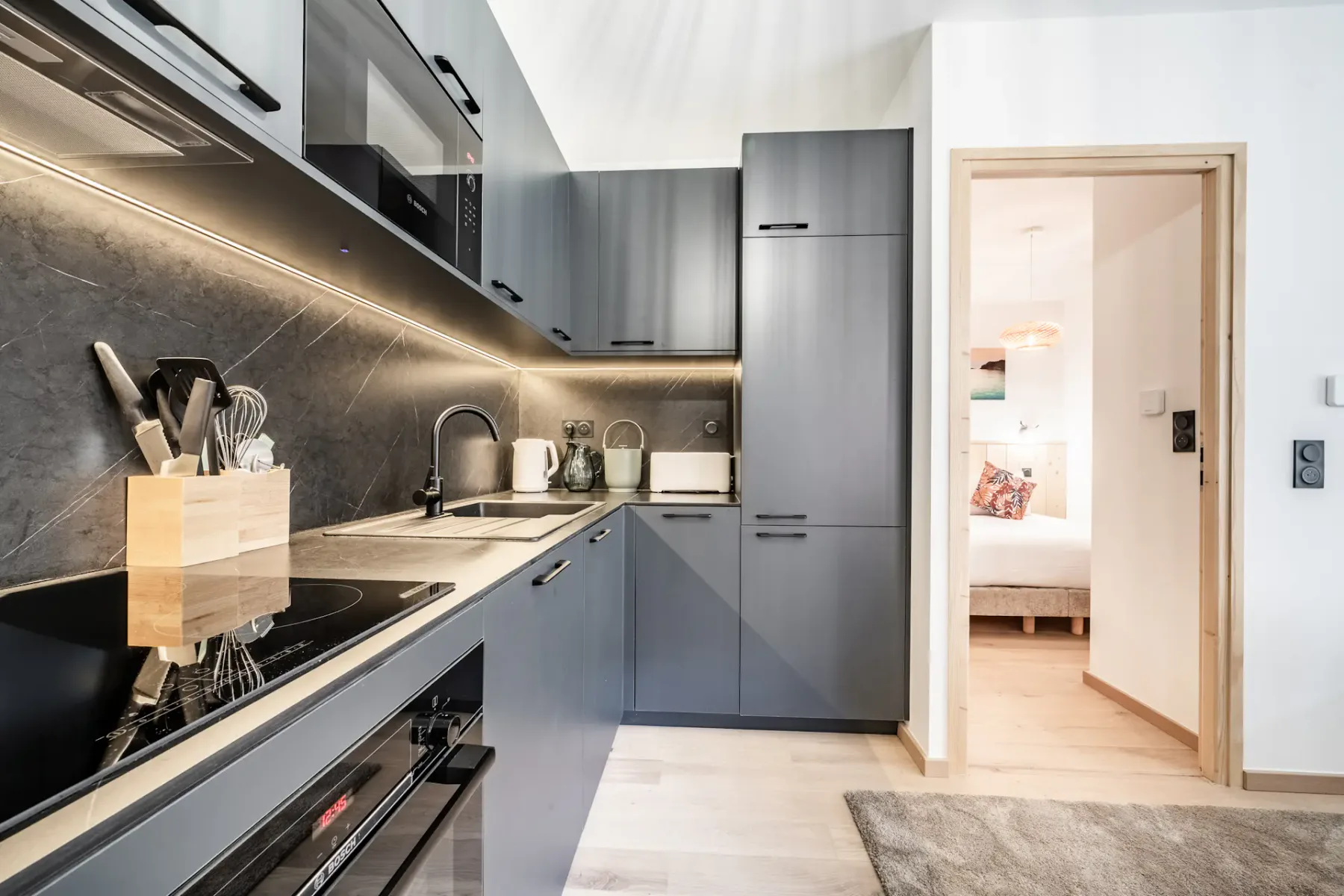 Modern kitchen with gray cabinets, built-in appliances, and a knife block on the counter. Doorway leads to a brightly lit bedroom.