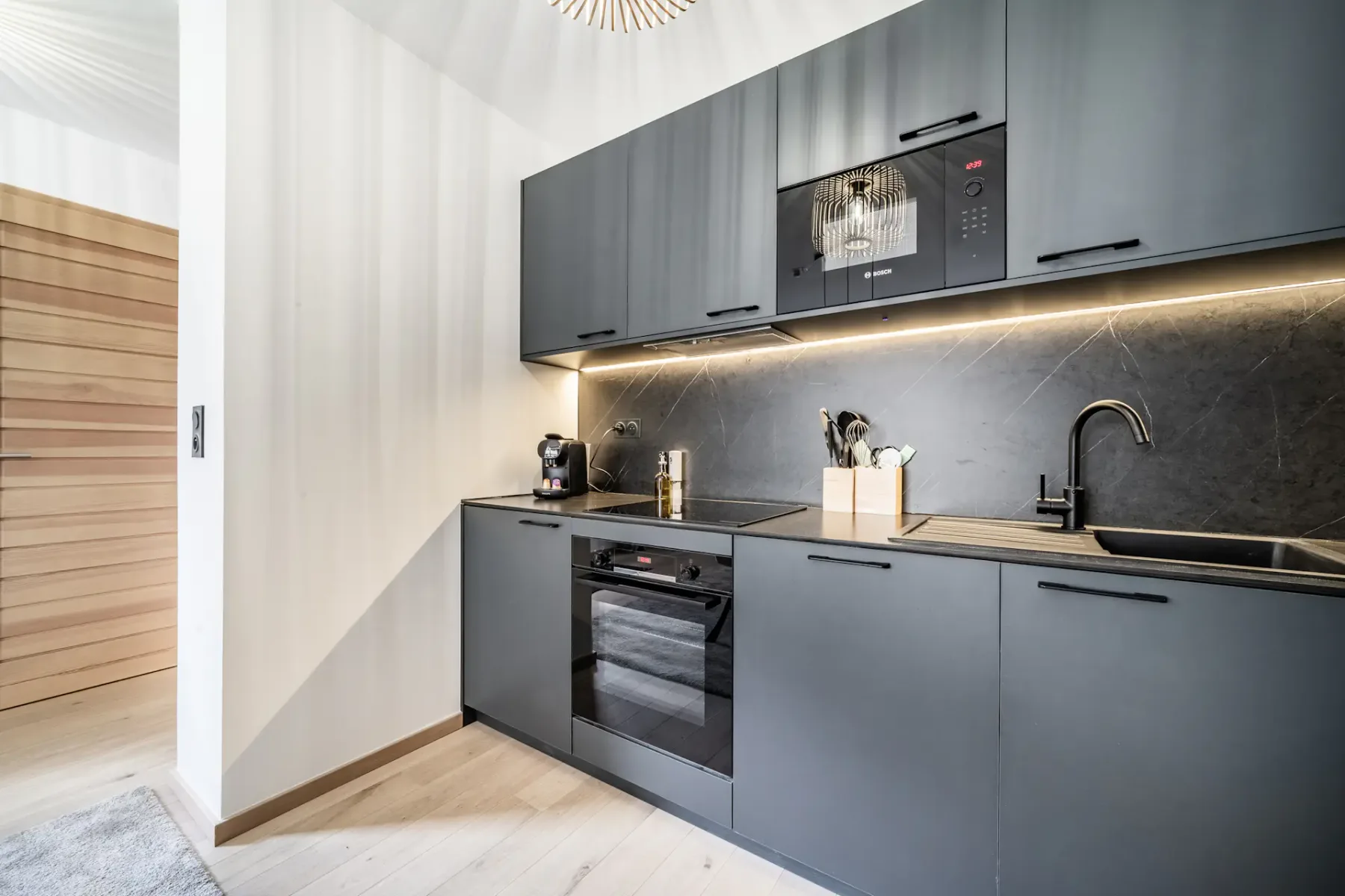 Modern kitchen with gray cabinets, built-in appliances, and a dark backsplash.
