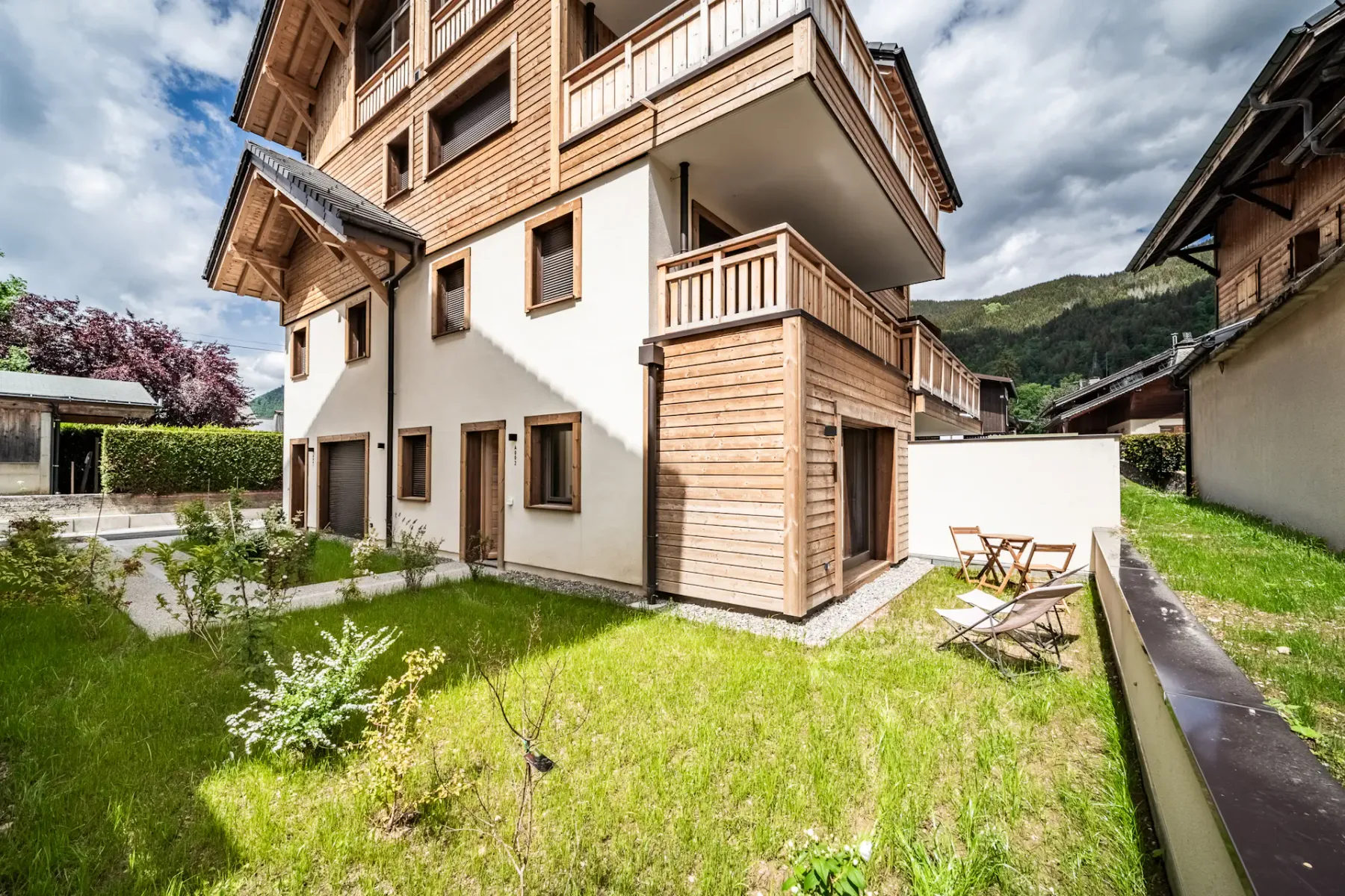 Modern building courtyard with greenery, wooden accents, open door, and mountain view under a partly cloudy blue sky.