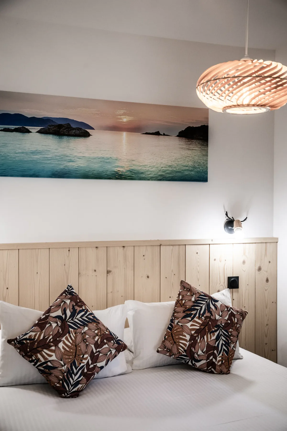 A bedroom with patterned pillows on a bed beneath a large coastal landscape print.