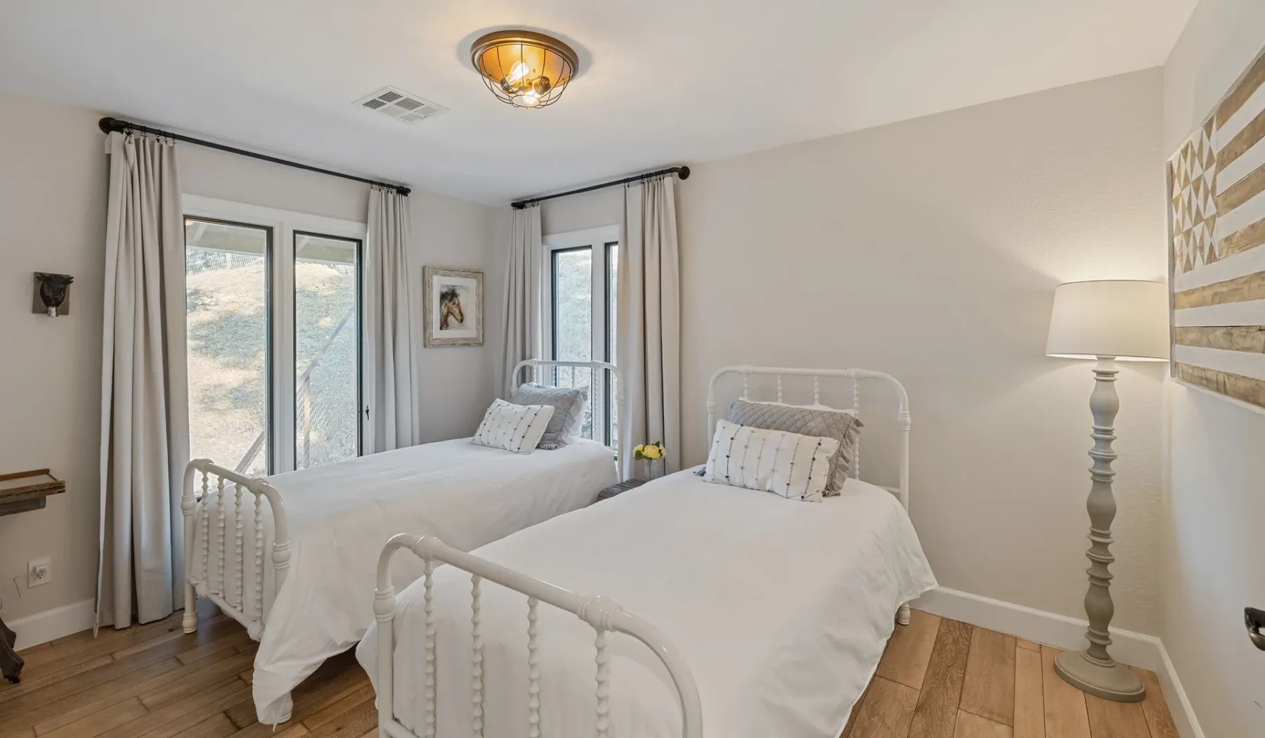Un dormitorio luminoso con dos camas junto a ventanas altas con cortinas.