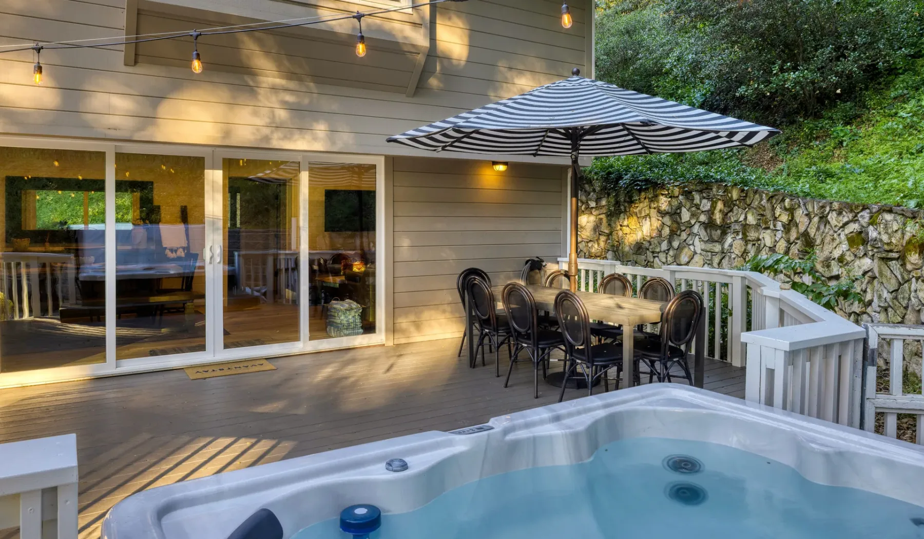 Una terraza exterior con una bañera de hidromasaje, mesa de comedor y sombrilla a rayas.
