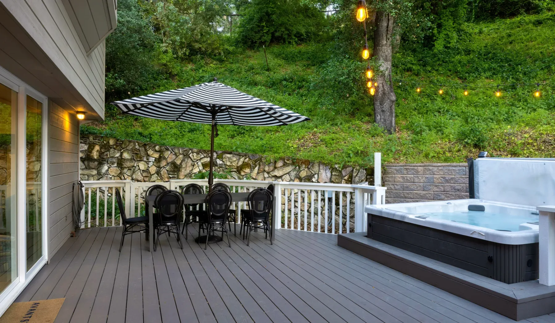 Una terraza exterior tiene una mesa de comedor bajo una sombrilla a rayas y una bañera de hidromasaje.