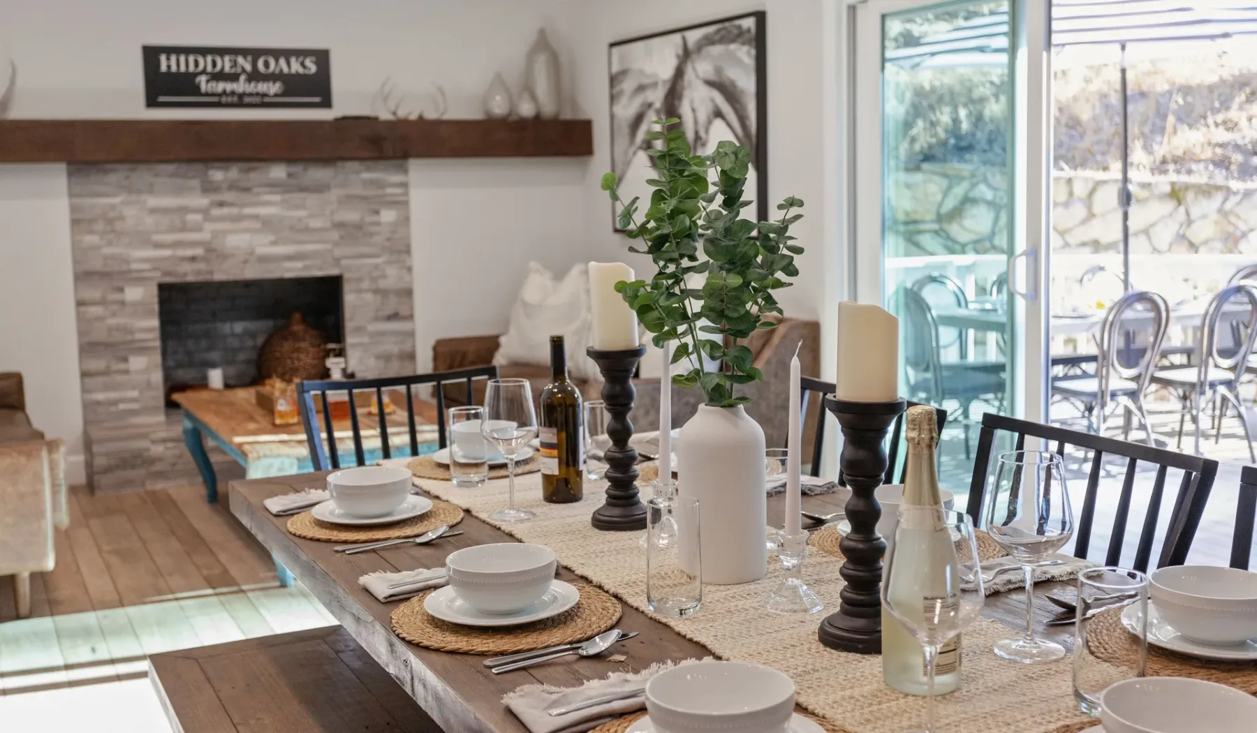 Una mesa de comedor está puesta junto a una sala de estar con chimenea y grandes ventanas.