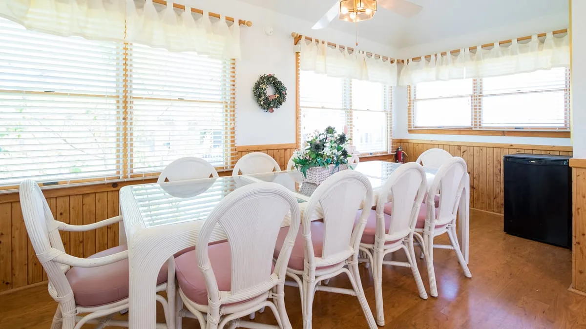 A bright dining room with a long table and white chairs beside large windows.