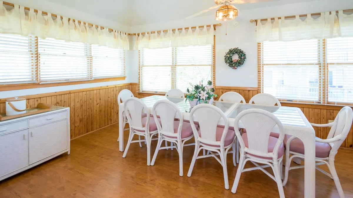 Un comedor luminoso con una mesa larga, sillas blancas y ventanas en las paredes.