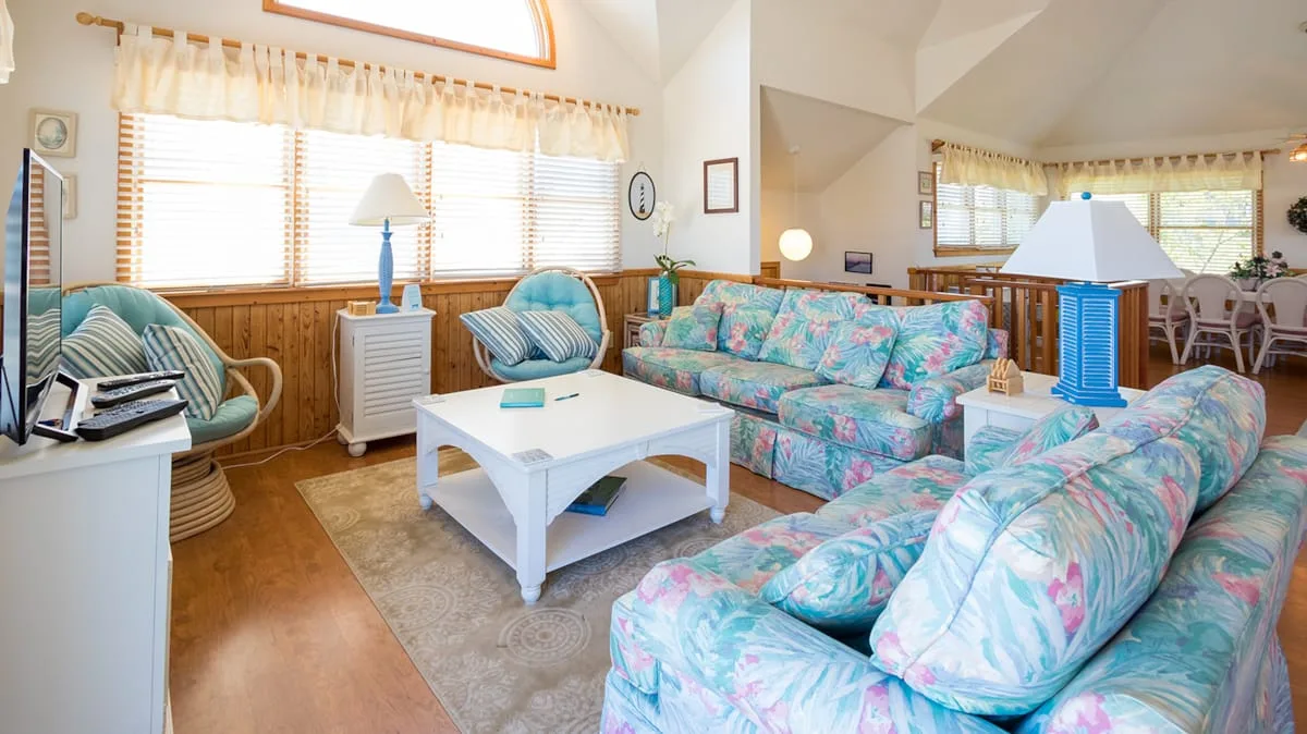 Una sala luminosa con asientos florales, una mesa de centro blanca y ventanas grandes.