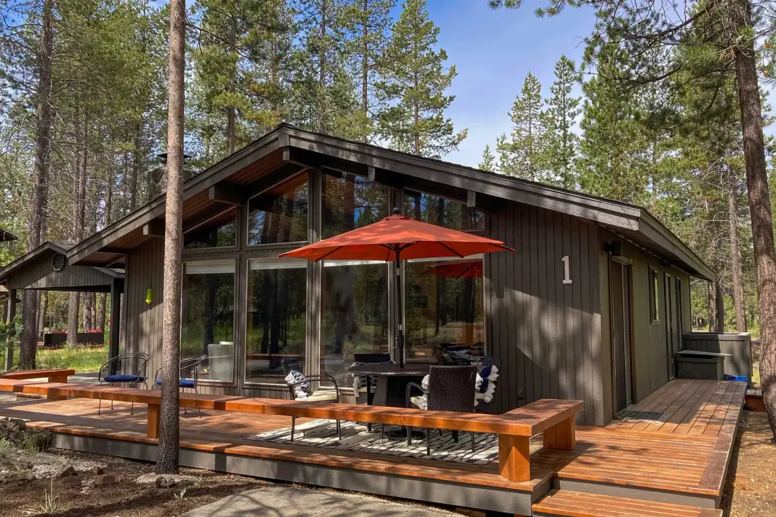 Una cabaña moderna con una amplia terraza de madera y una sombrilla naranja entre los árboles.