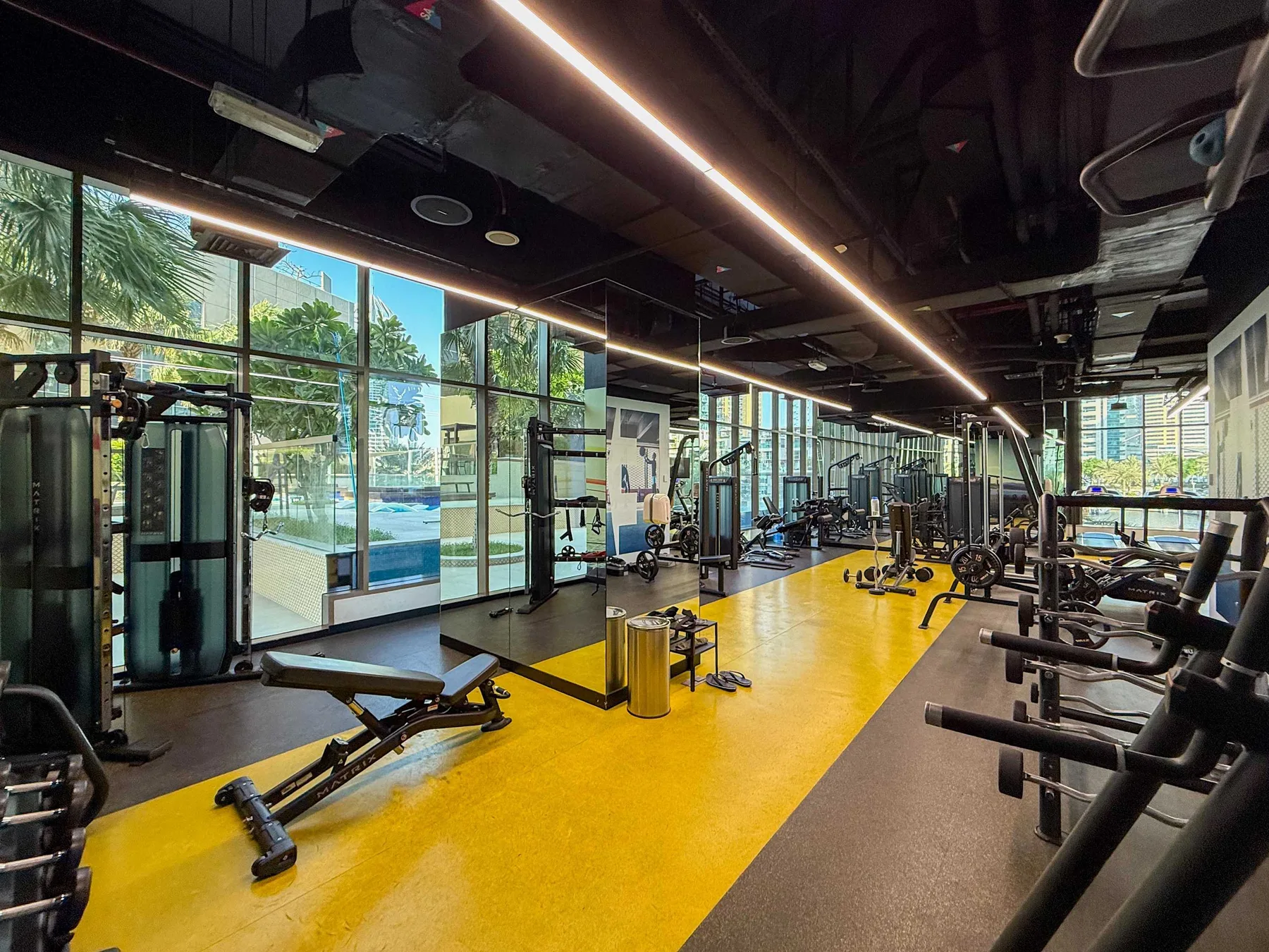 Un gimnasio con máquinas de ejercicio y bancos junto a grandes ventanales.