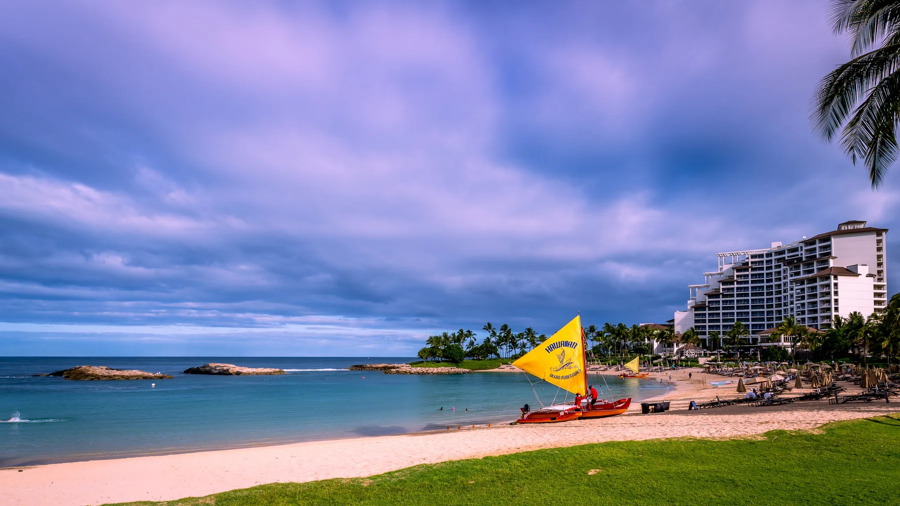 Una playa tropical junto a aguas tranquilas, con un edificio turístico a lo lejos.