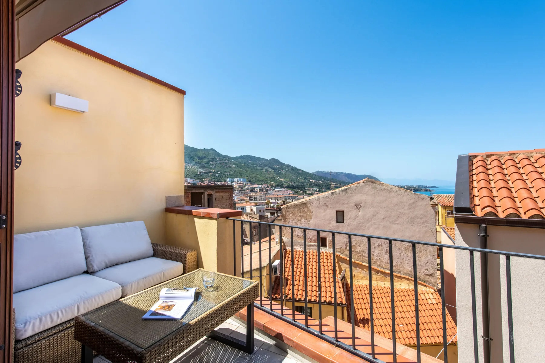 A small balcony seating area overlooks rooftops, hills, and the sea.