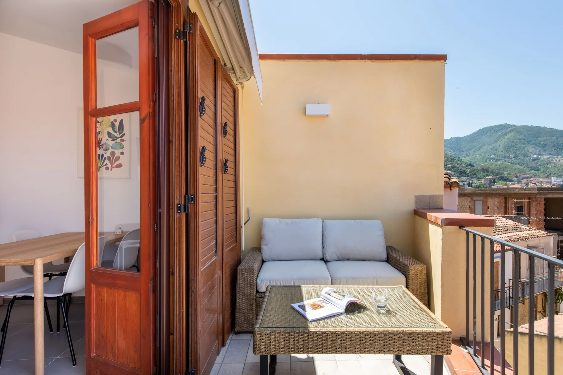 A sunny terrace has a small seating area beside open wooden doors.