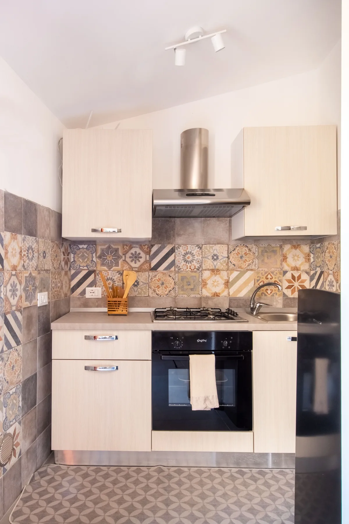 A compact kitchen with pale cabinets, a cooktop, and patterned tile.