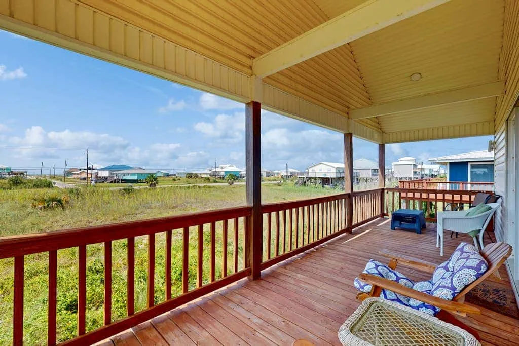 A covered porch with wooden railings and outdoor seating overlooks an open grassy area.