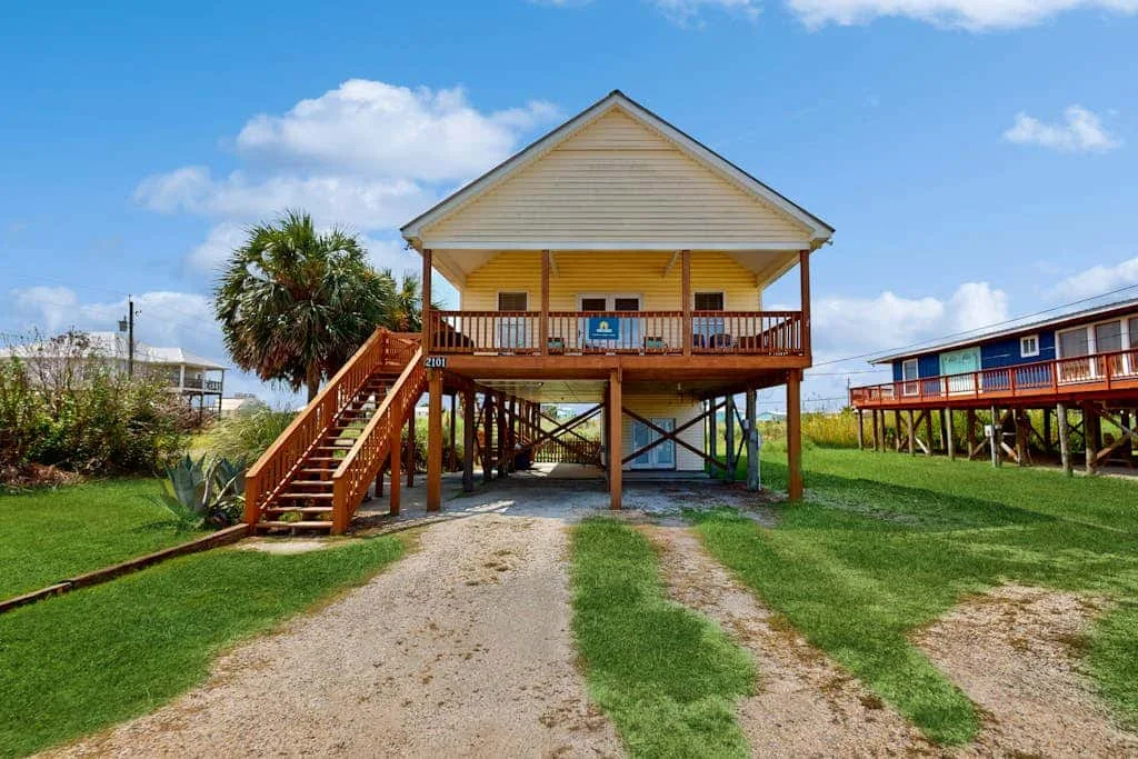 A raised house with a front porch stands above a grassy yard.