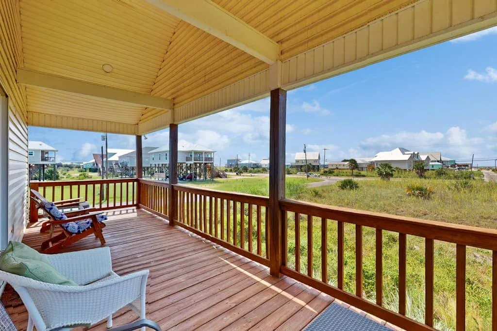 A covered wooden deck overlooks a grassy field and nearby buildings.
